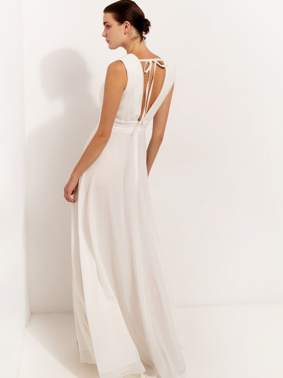 Λευκό μακρύ φόρεμα με σκίσιμο, αμάνικο, V ντεκολτέ, ελαφρύ ύφασμα, σούρα στην μέση, λεπτομέρεια άνοιγμα με φιόγκο στην πλάτη και κλείσιμο με φερμουάρ, εσωτερική υφασμάτινη επένδυση, κανονική εφαρμογή. Ελληνικής ραφής