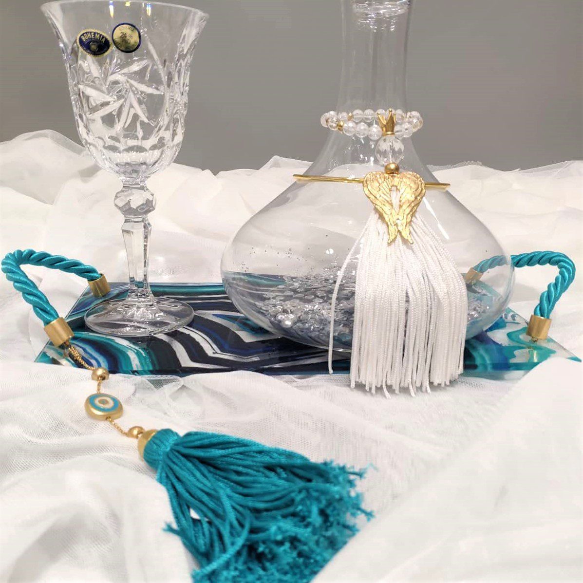 Σετ κουμπάρου σε κρύσταλλο Βοημίας με επίχρυσο στολισμό Tresento. Το σετ περιλαμβάνει περίτεχνο δίσκο με σμάλτο και πλεξιγκλάς, ιδιαίτερη μποτίλια κρασιού και ποτήρι από κρύσταλλο Βοημίας, διακοσμημένο με επιχρυσωμένο 24 καρατίων στολίδι Tresento με φτερά αγγέλου, με πέταλο, γαλάζια φούντα και μεταξωτό κορδόνι. Όλα είναι χειροποίητα και κατασκευασμένα στην Ελλάδα.